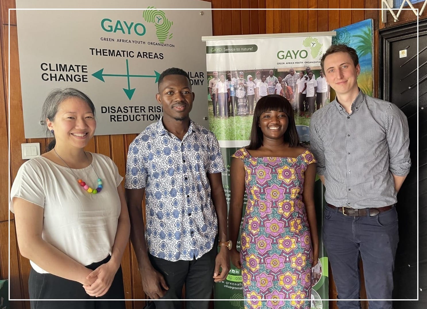 Four people standing in front of the Green Africa Youth Organization's office in Accra, Ghana. A sign shows GAYO's thematic areas of climate change, circular economy, and disaster risk reduction.