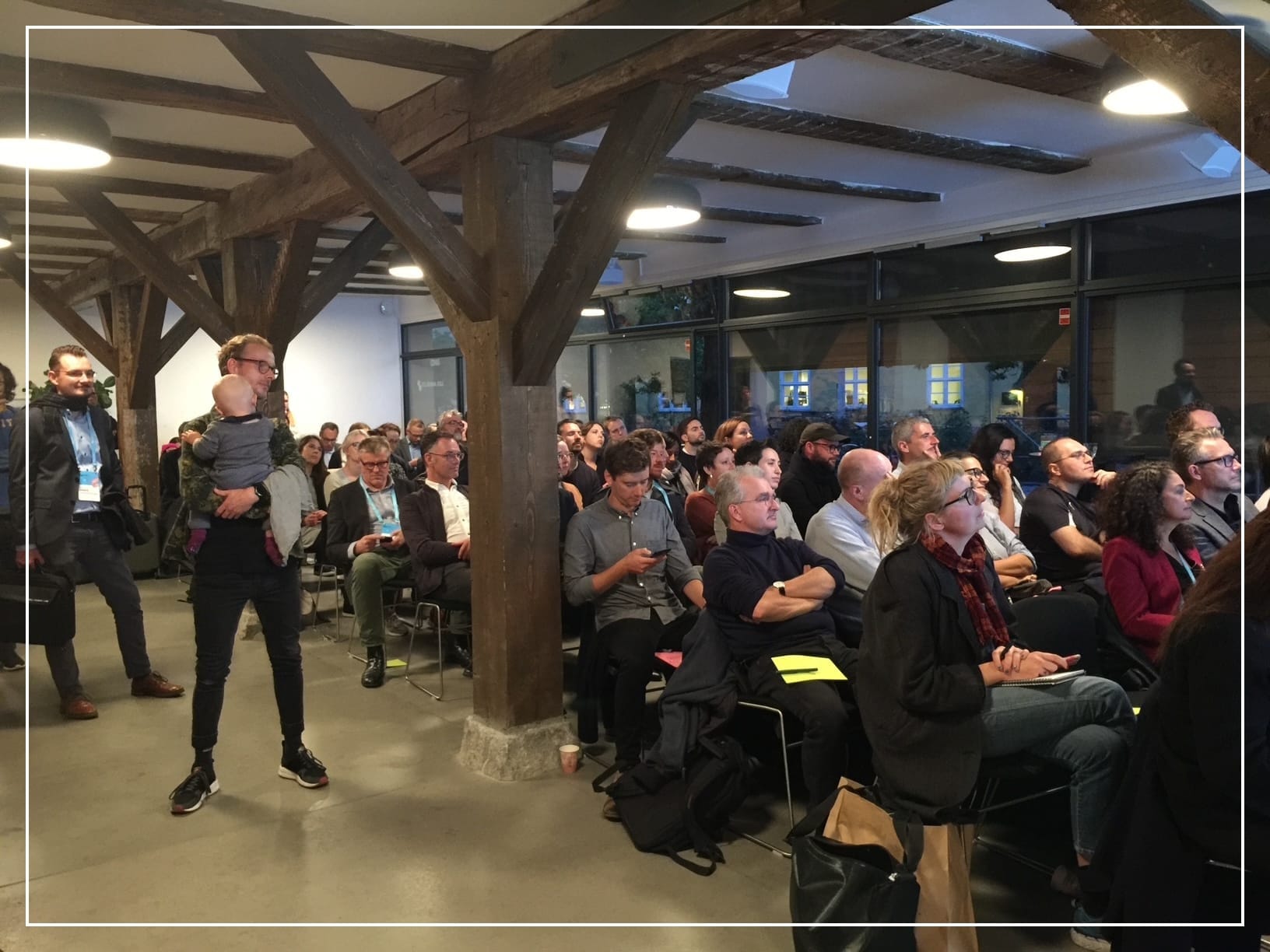Audience at a side session at the C40 Mayors' Summit in Copenhagen in 2019, seated in a wooden-beamed hall, listening intently. A man stands near the front holding a baby, adding warmth and everyday life to the scene.