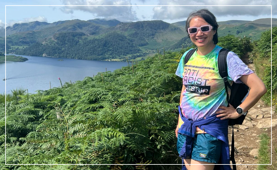 Woman in tie-dye t-shirt with Anti Racist Cumbria slogan, standing in a trail above a blue lake amidst green bracken.