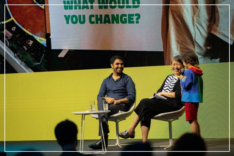 A man and a woman seated on a conference stage, smiling as they listen to a child speaking, in front of a giant image of a sign ending in "...what would you change?"