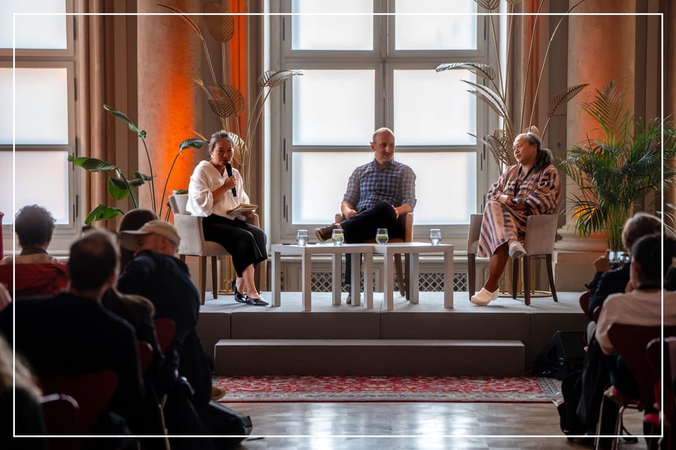 Two women and a man seated on an elevated platform at a conference side session, ample natural light and potted plants.
