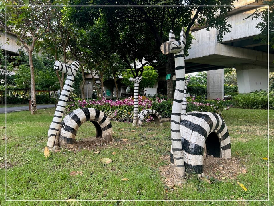 Zebra sculptures made of tires and logs painted black & white, surrounded by green grass, orchids, and trees under a highway in Bangkok.